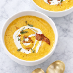 Creamy butternut squash soup topped with bacon, sage, and a swirl of cream in white bowls on a marble countertop