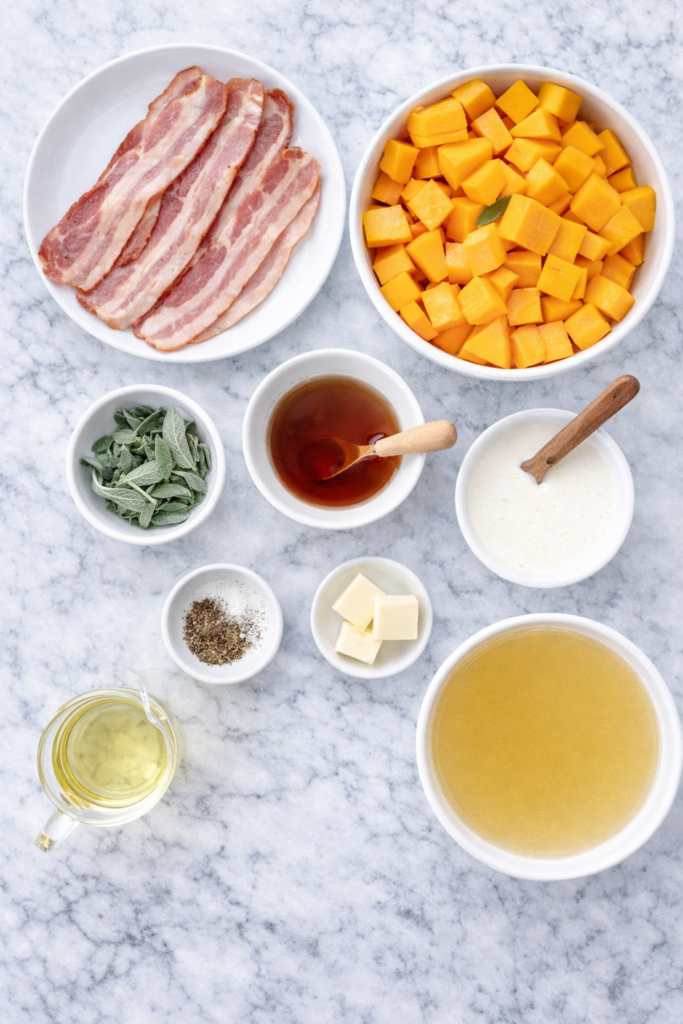 Flat lay of ingredients for butternut squash soup including squash, bacon, sage, maple syrup, cream, butter, and broth on a marble countertop