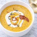 Creamy butternut squash soup with bacon, sage, and a swirl of cream in a white bowl on a marble countertop