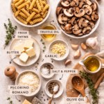 Flat lay of rotini pasta ingredients including baby bella mushrooms, truffle oil, garlic, butter, fresh thyme, and Pecorino Romano on a light neutral surface.