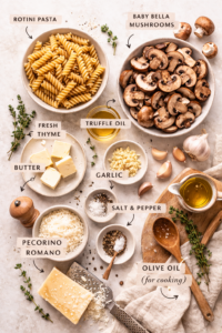 Flat lay of rotini pasta ingredients including baby bella mushrooms, truffle oil, garlic, butter, fresh thyme, and Pecorino Romano on a light neutral surface.