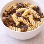 Vertical bowl of rotini pasta with sautéed mushrooms and truffle oil in a white ceramic bowl on a light neutral background.