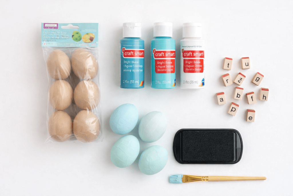 Flat lay of DIY Easter egg decorating supplies including paper mache eggs, pastel blue painted eggs, acrylic craft paint, letter stamps, ink pad, and paintbrush on a clean white background.