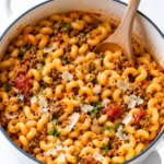 Johnny Marzetti pasta in a white enamel Dutch oven with a wooden spoon, topped with Parmesan cheese and fresh herbs on a white marble countertop.