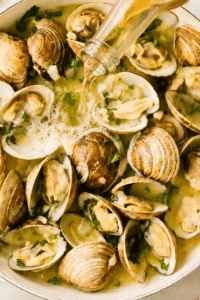 Pouring pale lager into littleneck clams with garlic and butter in a white enamel pan