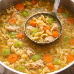 Homemade chicken noodle soup simmering in a stainless steel pot with egg noodles, carrots, celery, and shredded chicken in golden broth.