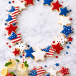 Patriotic red, white, and blue sugar cookie wreath arranged on a white marble background with loose star cookies and glasses of lemonade.