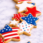 Close-up of patriotic red, white, and blue star-shaped sugar cookies decorated with simple stripes, stars, and polka dots on a marble surface.