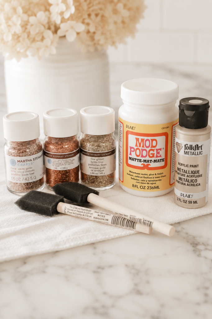 Supplies for making DIY glitter pumpkins including Mod Podge, glitter, metallic paint, and foam brushes on a white marble countertop.