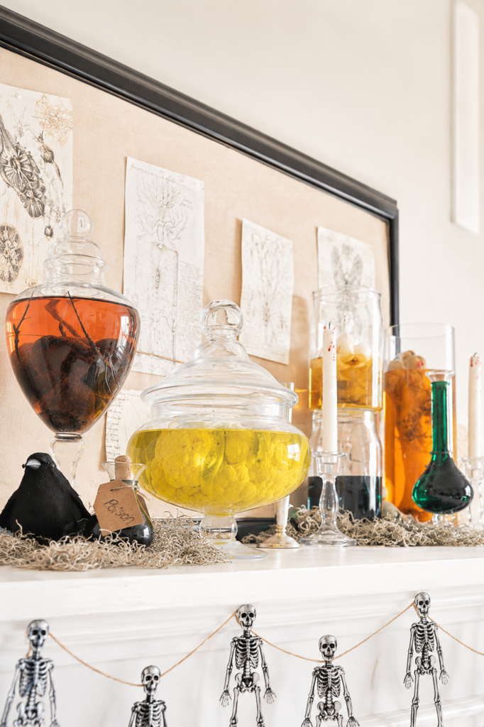 DIY Halloween specimen jars filled with colorful liquids displayed on a bright farmhouse-style mad scientist mantel with apothecary bottles, anatomy sketches, and skeleton garland.