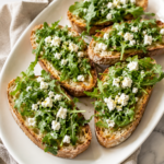 Easy arugula feta crostini topped with fresh lemon garlic arugula and crumbled feta cheese on a white platter.