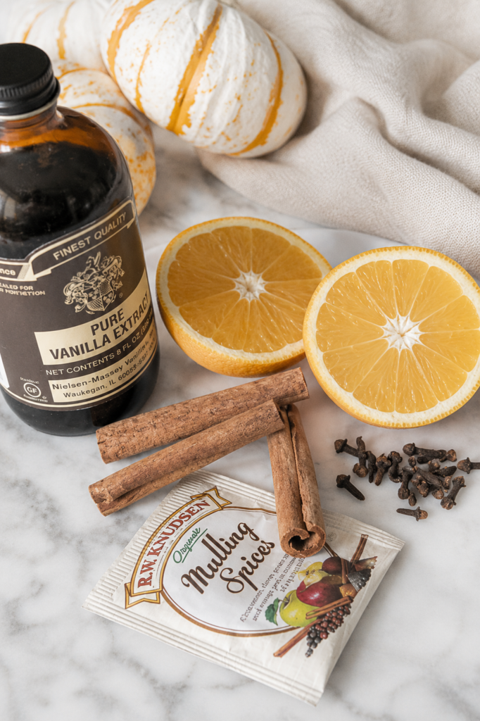Flat lay of ingredients for a fall simmer pot recipe including vanilla extract, cinnamon sticks, whole cloves, orange slices, and mulling spices on a white marble countertop with a neutral linen cloth.