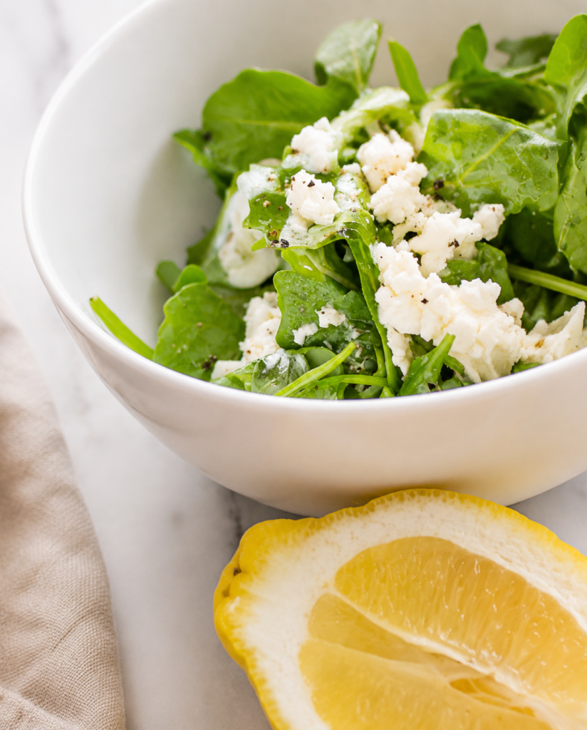 White bowl filled with lemon garlic arugula and crumbled feta cheese for arugula feta crostini.