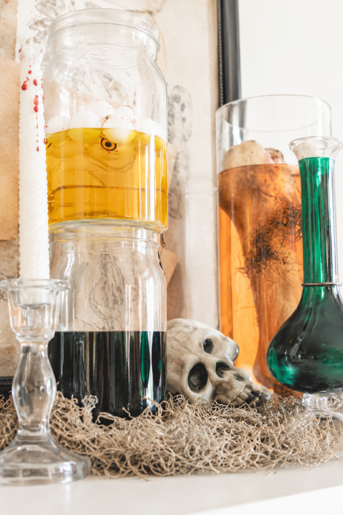 Close-up of Halloween specimen jars filled with eyeballs and colorful potions alongside vintage glass bottles, a skull, and spooky laboratory decor on a bright farmhouse mantel.
