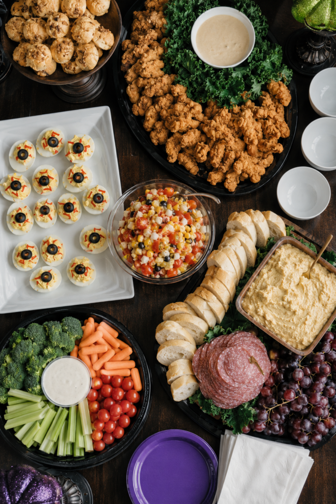 Overhead view of a witch-themed Halloween party buffet with deviled egg eyeballs, chicken fingers, vegetables, dip, bread, grapes and purple plates