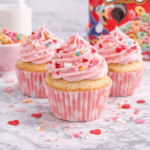 Homemade cereal milk Valentine’s cupcakes topped with pink buttercream frosting and heart sprinkles with a box of fruity cereal in the background