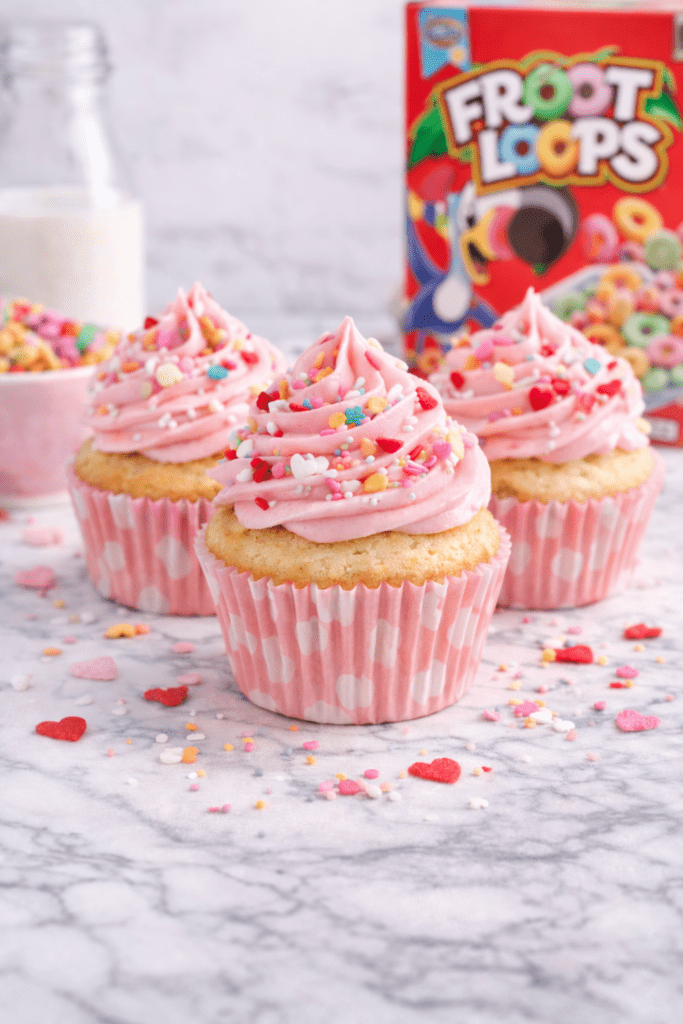 Homemade cereal milk Valentine’s cupcakes topped with pink buttercream frosting and heart sprinkles with a box of fruity cereal in the background
