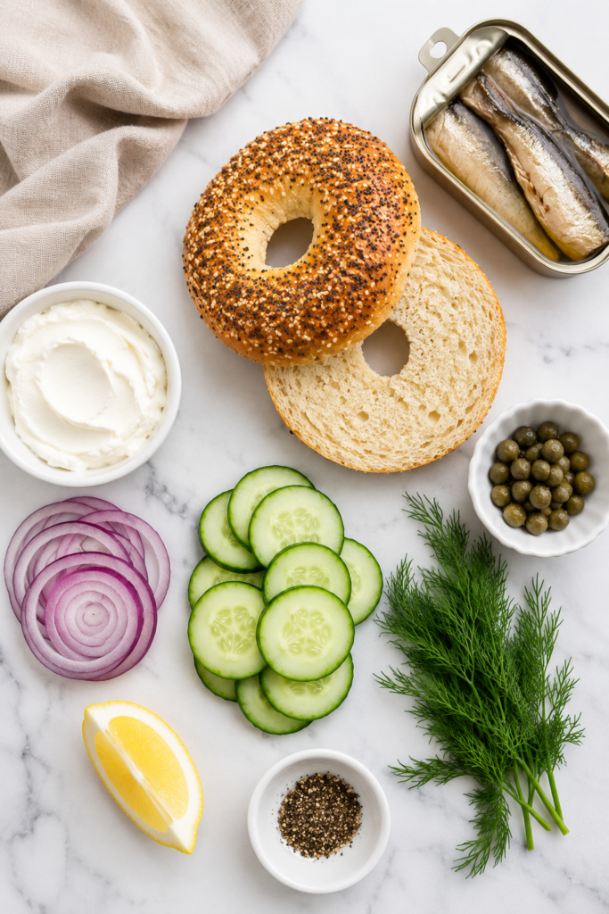 Flat lay of ingredients for a sardine cream cheese breakfast bagel including an everything bagel, sardines, cream cheese, cucumber slices, red onion, capers, fresh dill, lemon, and black pepper on a white marble countertop.