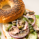 Sardine cream cheese breakfast bagel topped with cucumber, red onion, capers, and fresh dill on a white plate over a marble countertop.