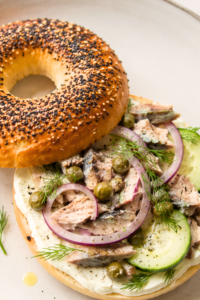 Sardine cream cheese breakfast bagel topped with cucumber, red onion, capers, and fresh dill on a white plate over a marble countertop.