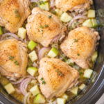 Seared chicken thighs arranged over diced green apples and sliced shallots with thyme and mustard broth in a black crockpot.