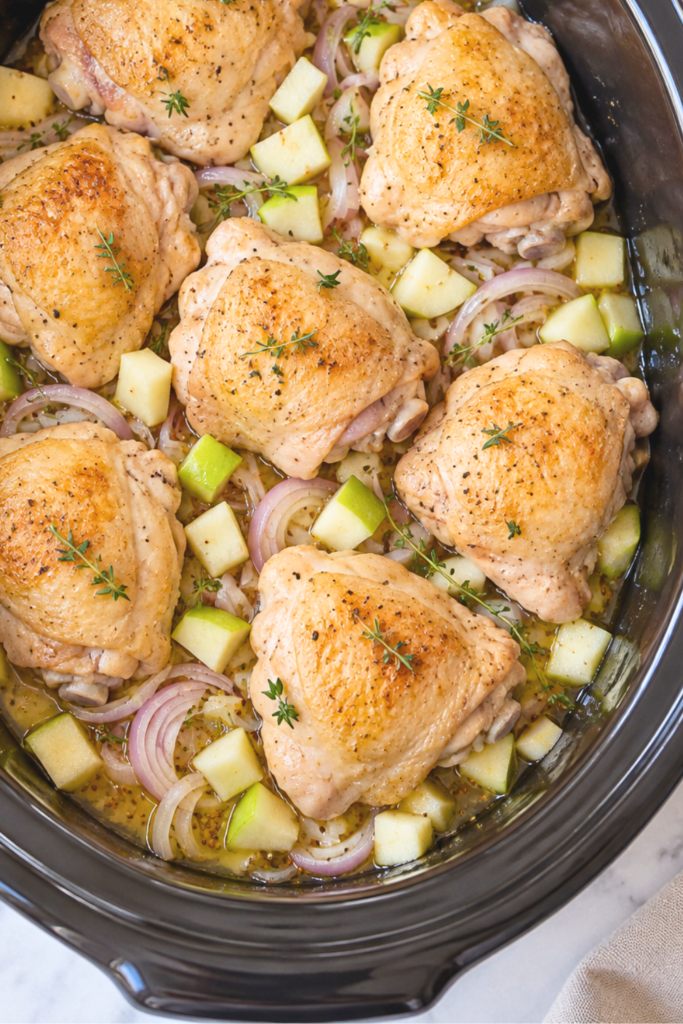 Seared chicken thighs arranged over diced green apples and sliced shallots with thyme and mustard broth in a black crockpot.