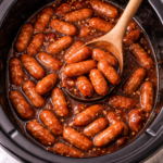 Slow cooker Little Smokies simmering in a sweet brown sugar sauce inside a crockpot with a wooden spoon.