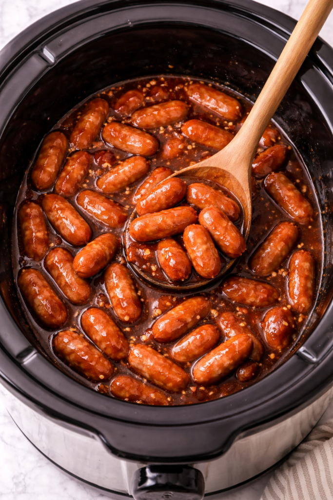 Slow cooker Little Smokies simmering in a sweet brown sugar sauce inside a crockpot with a wooden spoon.