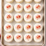Overhead view of strawberry almond petits fours lined up on a baking sheet with white chocolate glaze and strawberry topping