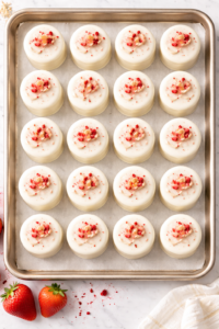 Overhead view of strawberry almond petits fours lined up on a baking sheet with white chocolate glaze and strawberry topping