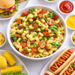 Birds eye view of BLT macaroni salad with bacon and tomatoes surrounded by hot dogs and hamburgers on a light marble table