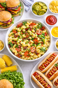 Birds eye view of BLT macaroni salad with bacon and tomatoes surrounded by hot dogs and hamburgers on a light marble table