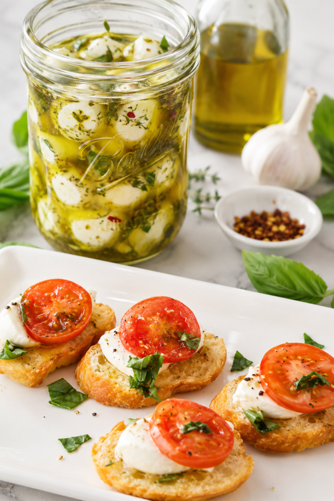 Marinated mozzarella crostini served on a white platter with fresh tomatoes and basil, with a jar of homemade herb-marinated mozzarella in the background.