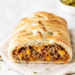 Golden bacon cheeseburger calzone sliced open to reveal seasoned ground beef, crispy bacon, and melted cheddar cheese on a marble countertop with a neutral linen cloth.