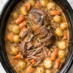 Creamy crock pot pot roast with tender shredded beef, baby potatoes, and carrots in a rich onion gravy inside a black slow cooker on a white marble countertop.