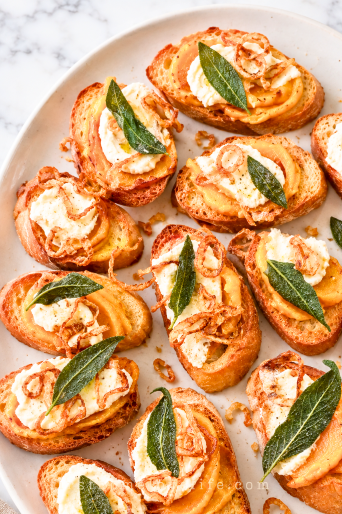 Squash butter crostini topped with ricotta, crispy shallots, and fried sage on a white platter over a marble countertop.