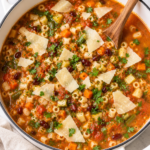 The best homemade minestrone soup in a white enamel Dutch oven topped with shaved Parmesan and fresh parsley on a marble countertop.