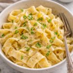 Bowl of homemade egg noodles tossed with melted butter and fresh parsley on a marble countertop.