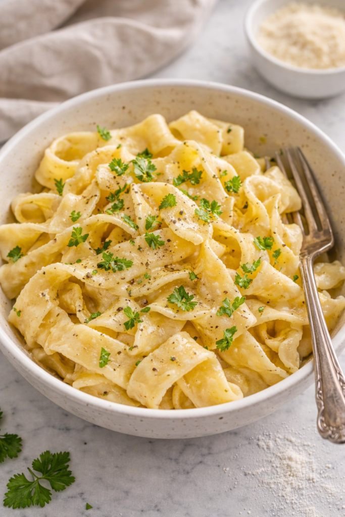 Bowl of homemade egg noodles tossed with melted butter and fresh parsley on a marble countertop.