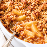 Homemade apple crisp with golden oat crumble and tender cinnamon apples in a white baking dish with a silver serving spoon.