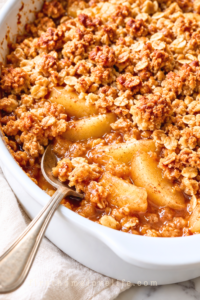 Homemade apple crisp with golden oat crumble and tender cinnamon apples in a white baking dish with a silver serving spoon.