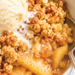 Warm apple crisp with golden oat crumble and vanilla ice cream in a white bowl with a silver spoon on a marble countertop.