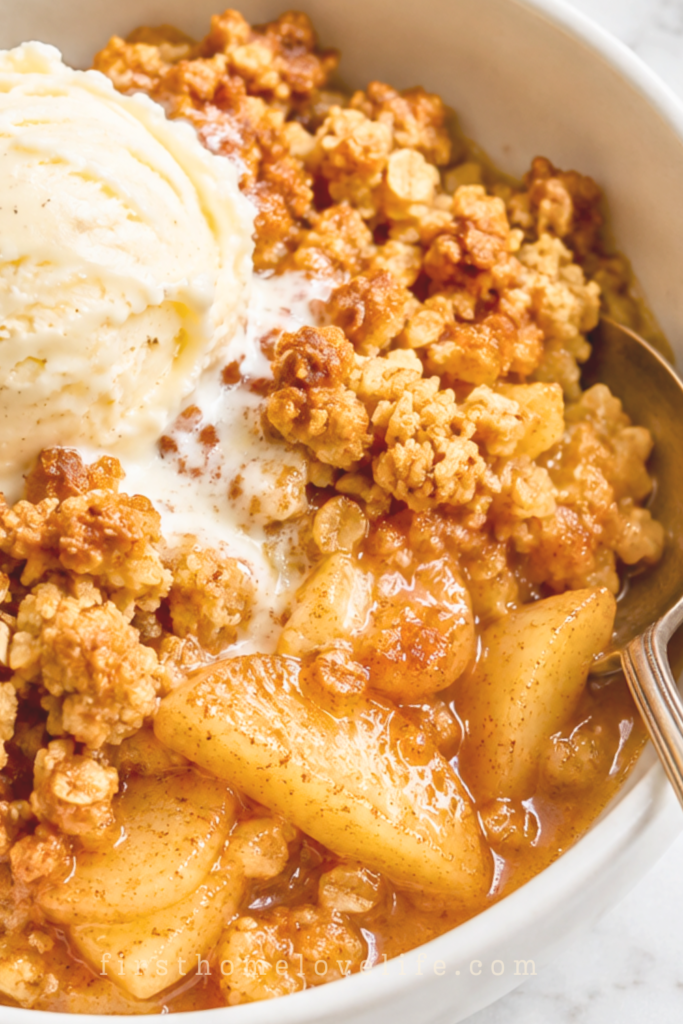 Warm apple crisp with golden oat crumble and vanilla ice cream in a white bowl with a silver spoon on a marble countertop.