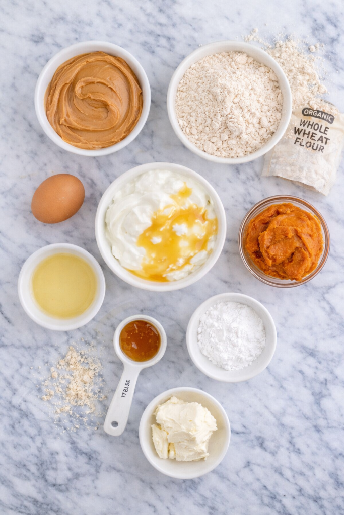 Flat lay of dog cupcake ingredients including peanut butter, whole wheat flour, pumpkin puree, Greek yogurt, honey, egg, and cream cheese on a marble countertop