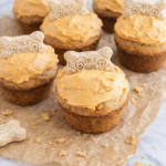 Homemade doggie cupcakes topped with pumpkin peanut butter frosting and dog bone treats, sitting on crinkled brown parchment paper on a marble countertop