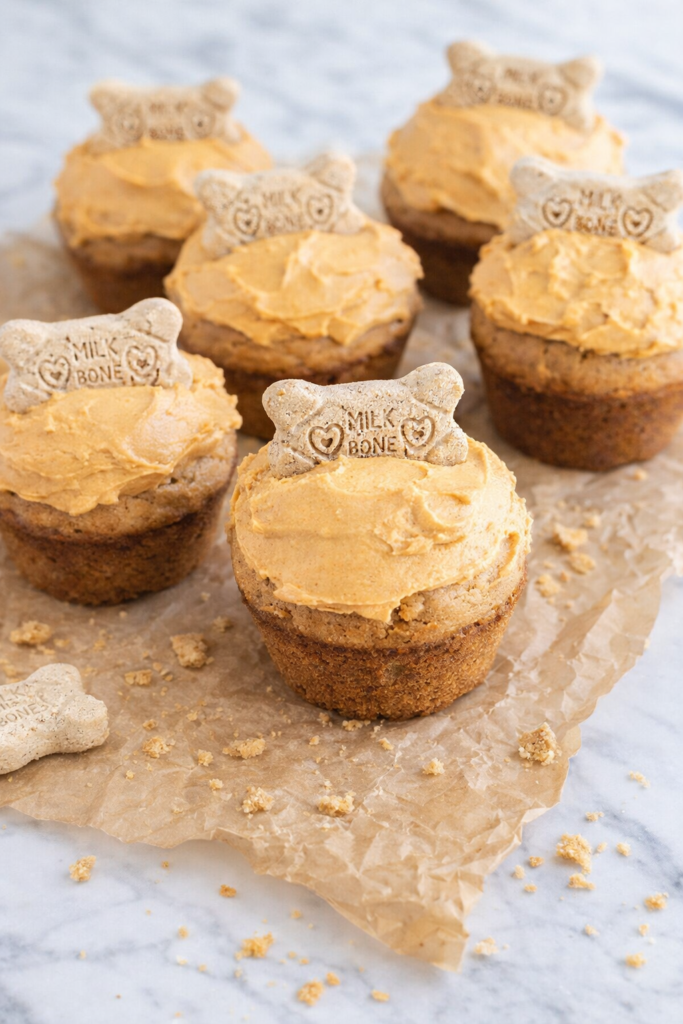 Homemade doggie cupcakes topped with pumpkin peanut butter frosting and dog bone treats, sitting on crinkled brown parchment paper on a marble countertop