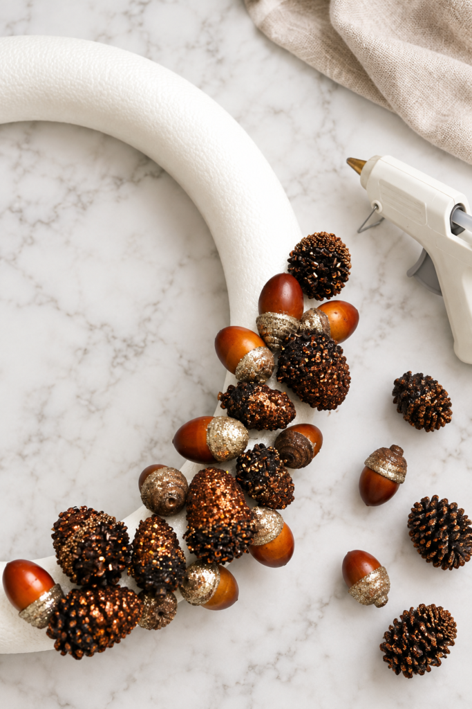 Partially assembled white foam wreath form with glittered gold pinecones and acorns, shown with a hot glue gun on a white marble countertop.