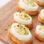 Deviled egg crostini on toasted baguette slices topped with creamy egg filling, fresh dill, and capers on a wooden board.