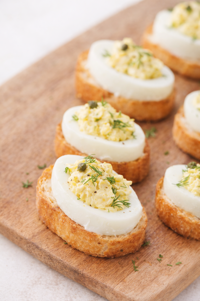 Deviled egg crostini on toasted baguette slices topped with creamy egg filling, fresh dill, and capers on a wooden board.