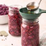 Pouring warm spiced vinegar brine into mason jars filled with shredded red cabbage using a funnel for water bath canning.